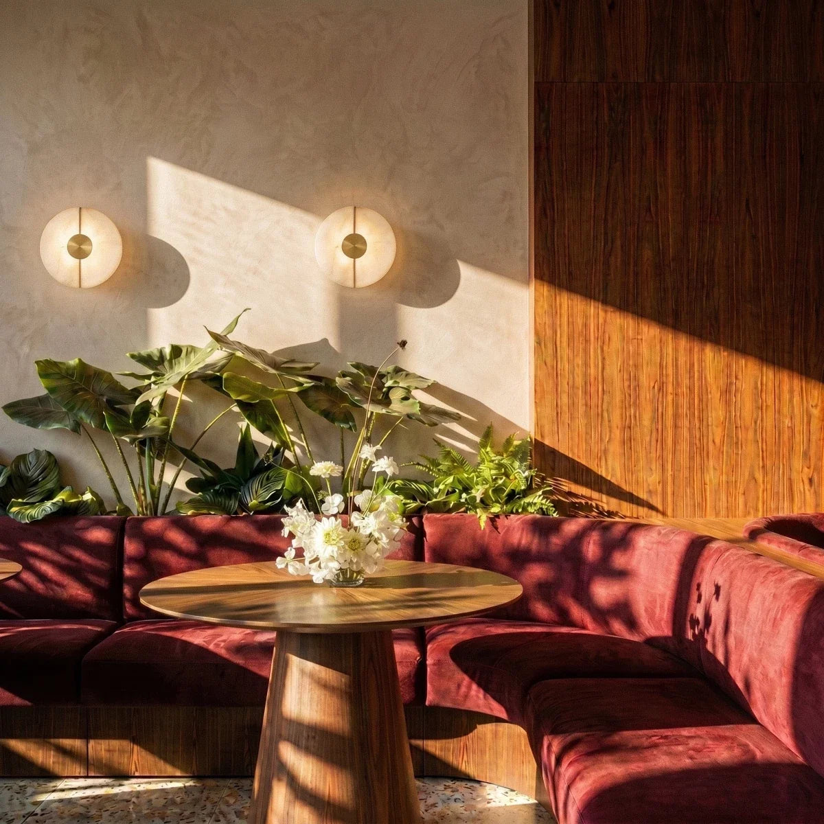 Two round Clérachic alabaster sconces cast warm shadows over a red velvet booth and lush plants at dusk.
