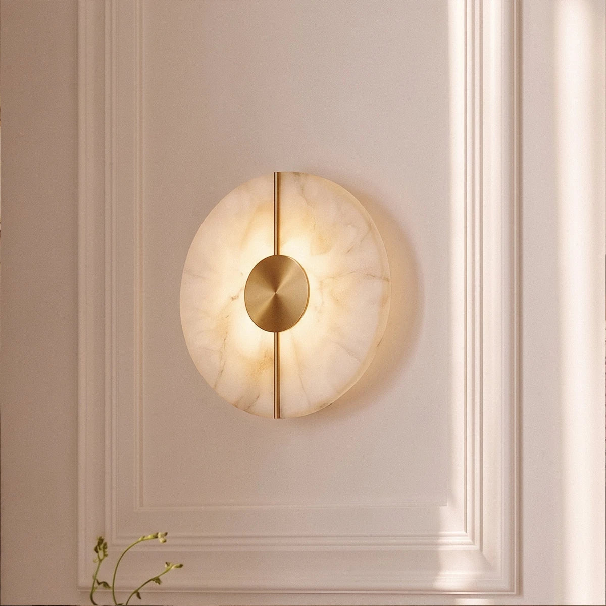 Set into a paneled wall, the round Clérachic alabaster sconce glows softly beside a trailing green plant stem.