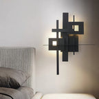 A lit Clérachic wall light with matt black overlapping panels and glowing square cutouts, on a pale wall beside a grey upholstered bed.