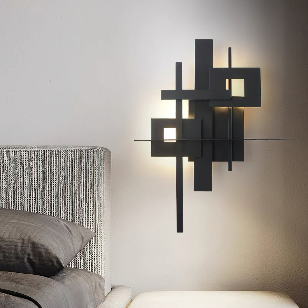 A lit Clérachic wall light with matt black overlapping panels and glowing square cutouts, on a pale wall beside a grey upholstered bed.