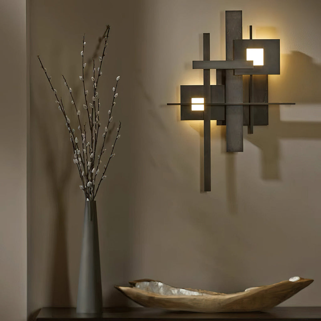 A Clérachic matt black wall light glowing from its square cutouts, on a warm taupe wall beside a tall vase of branches and a wooden bowl.