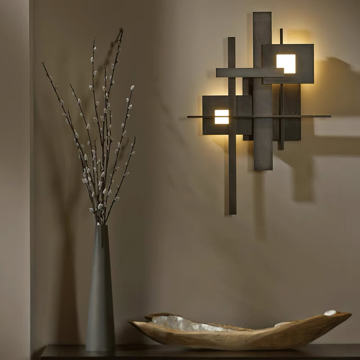 A Clérachic matt black wall light glowing from its square cutouts, on a warm taupe wall beside a tall vase of branches and a wooden bowl.