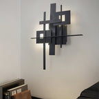 A Clérachic wall light of overlapping black panels with a long horizontal bar, casting a soft halo on a white wall above books and bedding.