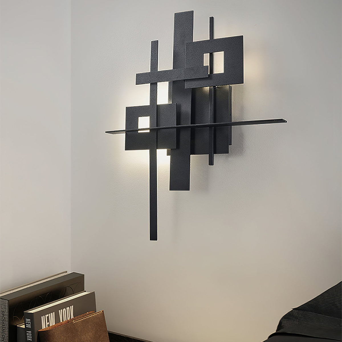 A Clérachic wall light of overlapping black panels with a long horizontal bar, casting a soft halo on a white wall above books and bedding.
