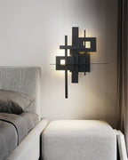 A Clérachic matt black wall light, lit, above a pale ottoman at the foot of a bed with grey bedding.