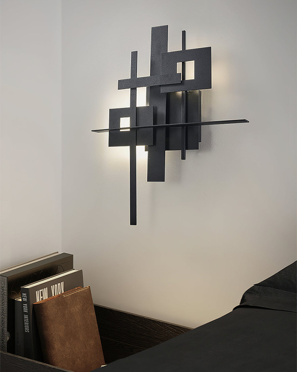 A Clérachic wall light of stacked black rectangles glowing softly on a white wall, with a stack of books and a dark bed below.