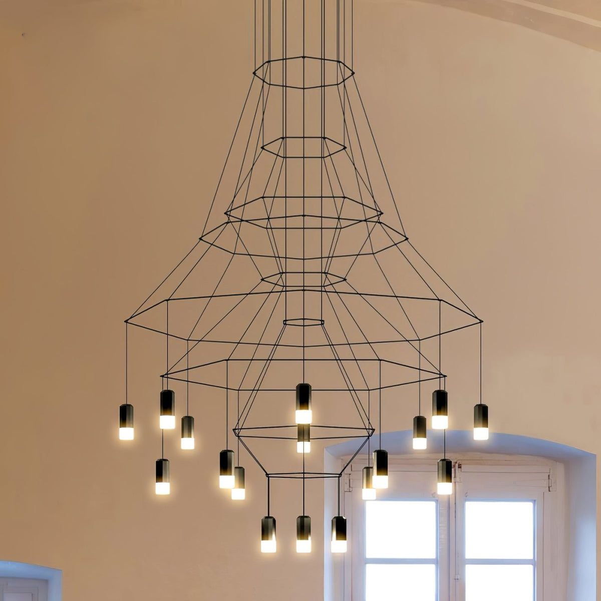 Under-view of the flared Clérachic wire chandelier, its fine black polygon frames narrowing to a lit lower cluster beside a window.