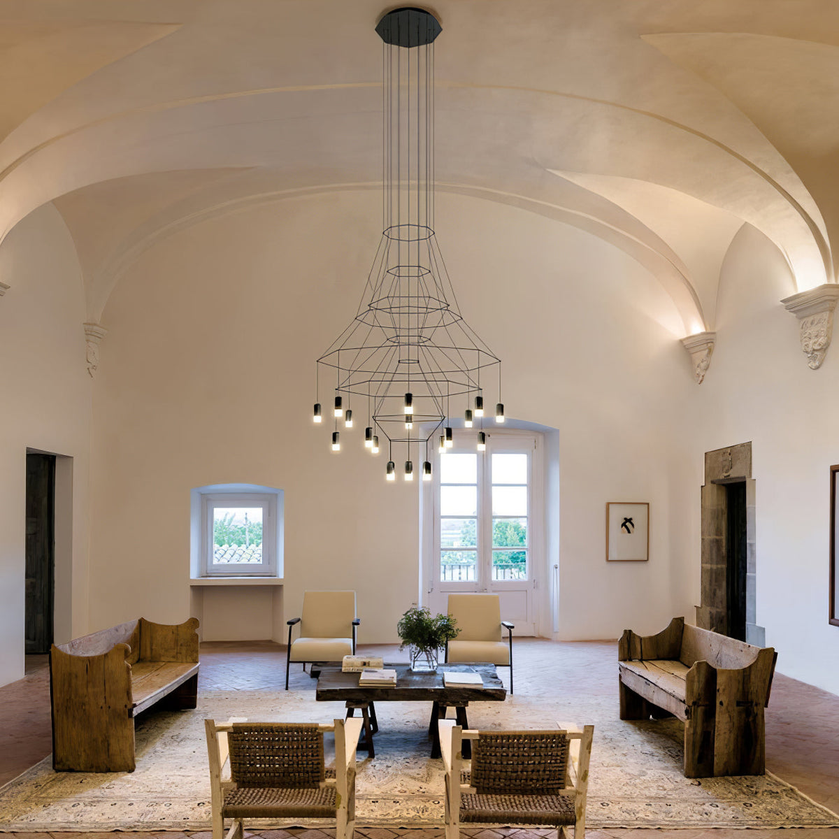 A Clérachic wire chandelier hangs from a long drop in a vaulted room, above a low table and rustic benches.