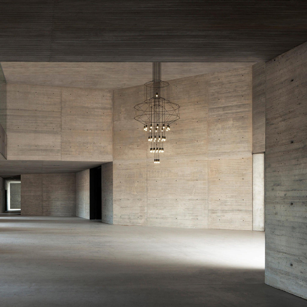 A Clérachic wire chandelier hung high in a raw concrete hall, with its LED drops lit and the floor below left bare.