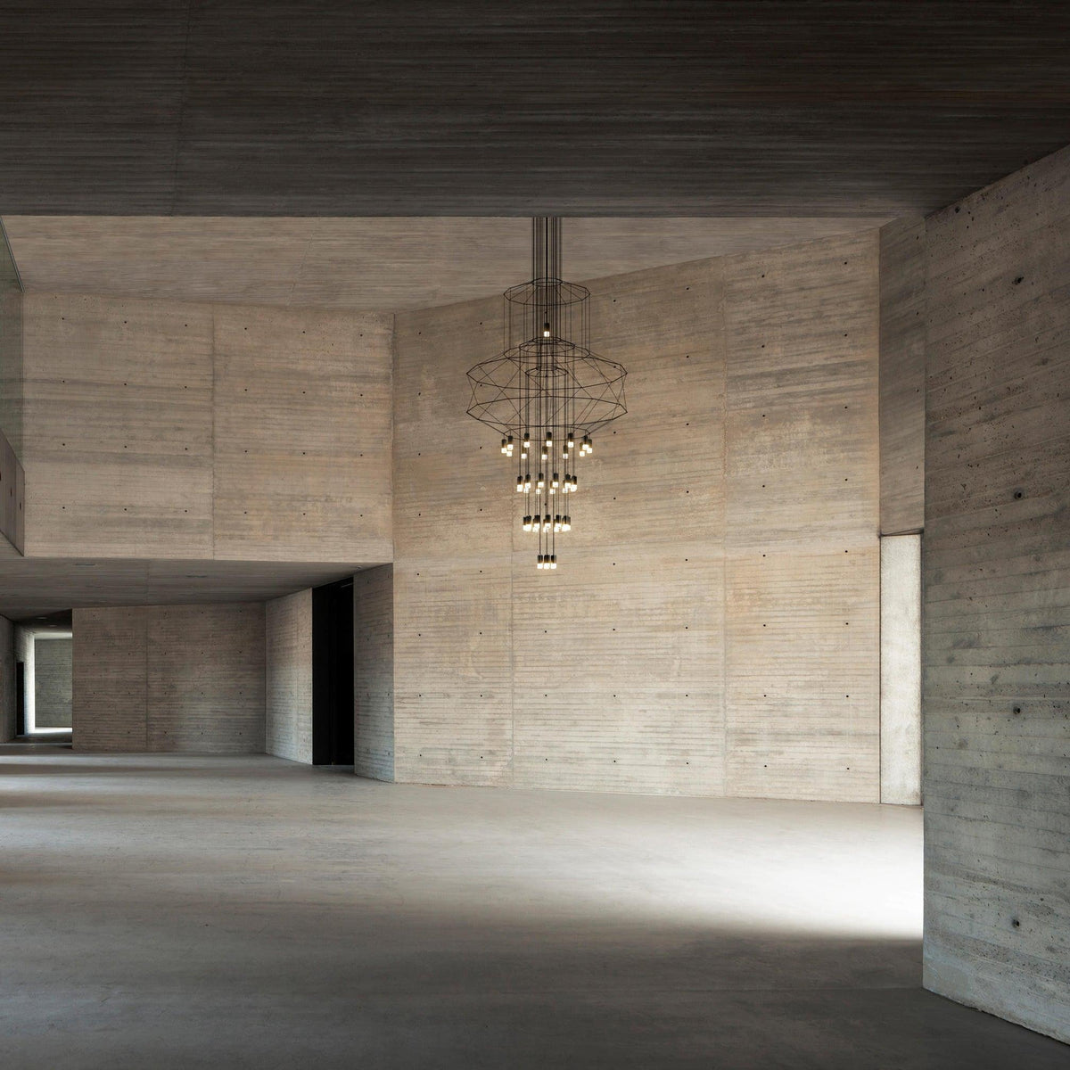 A Clérachic wire chandelier hung high in a raw concrete hall, with its LED drops lit and the floor below left bare.