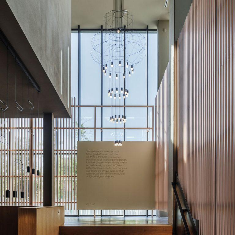 Tall Clérachic wire chandelier running down a glazed stairwell with timber slat walls, its LED drops lit one above another.