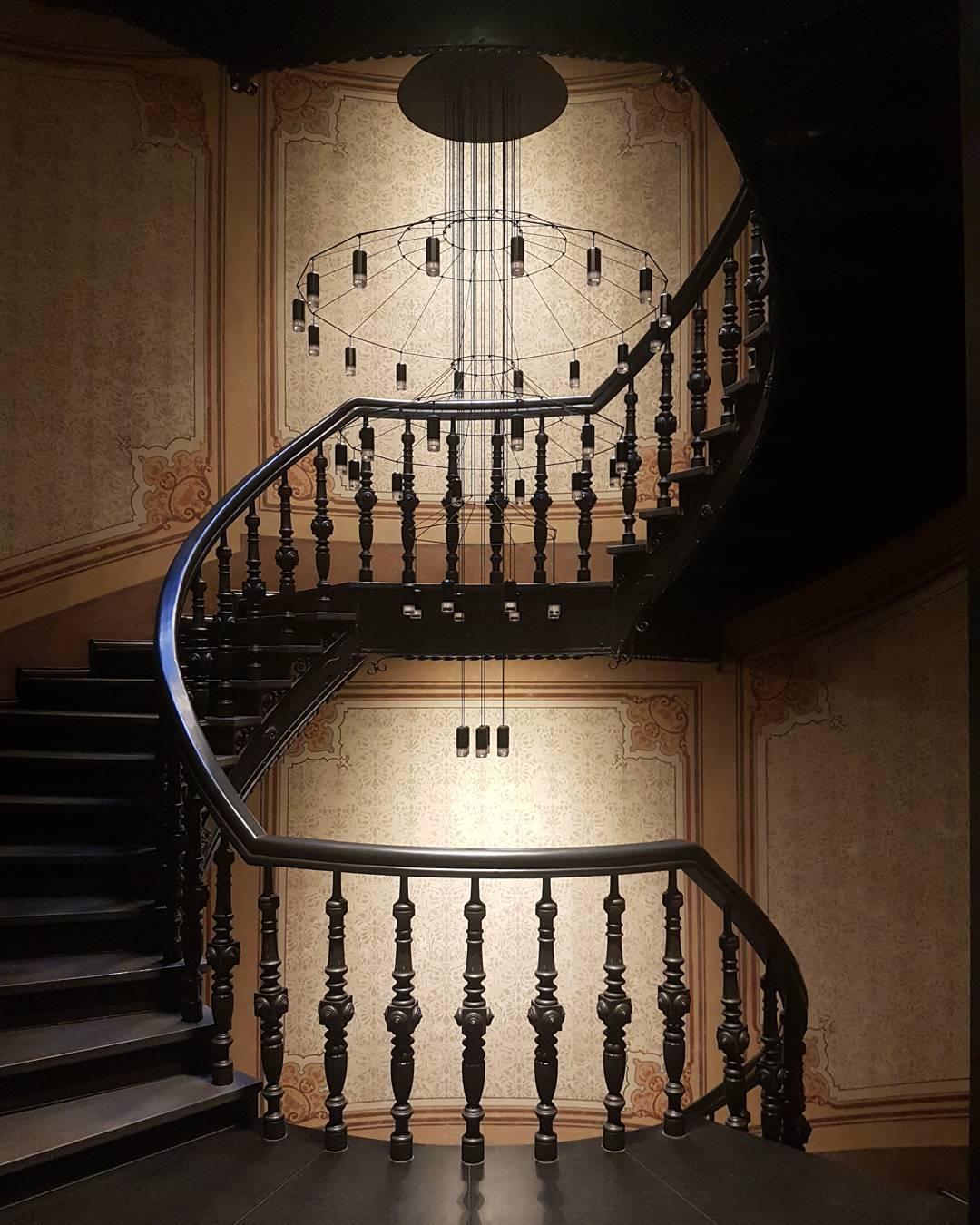 A Clérachic wire chandelier hangs in a dark spiral stairwell with carved black balusters and a warm-lit wall behind.