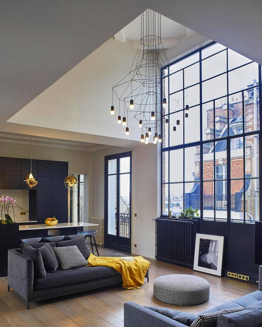 A Clérachic wire chandelier hanging in a double-height loft with a big steel-framed window, a grey sofa and a yellow throw.