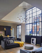 A Clérachic wire chandelier hanging in a double-height loft with a big steel-framed window, a grey sofa and a yellow throw.