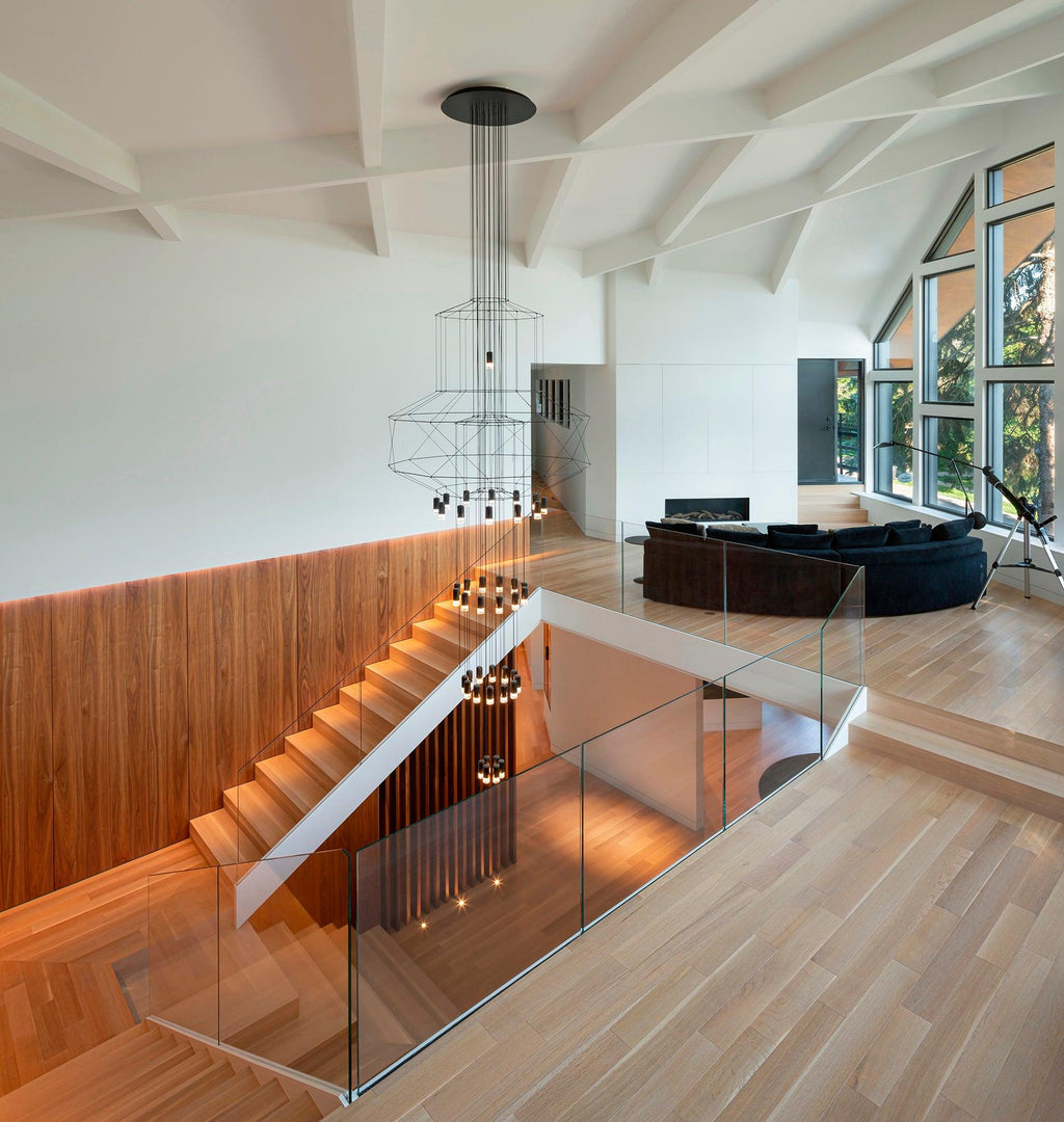 A Clérachic wire chandelier hangs above an open stair with a glass balustrade, a timber wall and white beams.