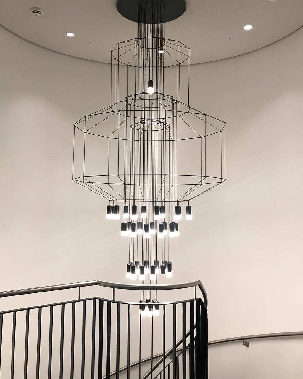 Clérachic wire chandelier seen from below in a round stairwell, with stacked frames, many LED drops and a black handrail.