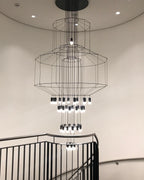 Clérachic wire chandelier seen from below in a round stairwell, with stacked frames, many LED drops and a black handrail.