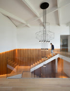 A person climbs a timber stair beneath a hanging Clérachic wire chandelier in a bright room with white beams.