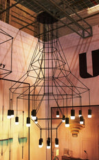 Looking up at a Clérachic wire chandelier with flared frames and lit LED drops in a warm-lit exhibition-style space.