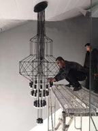 Two people fit a stacked-frame Clérachic wire chandelier from a scaffold platform, with many LED drops already hung.