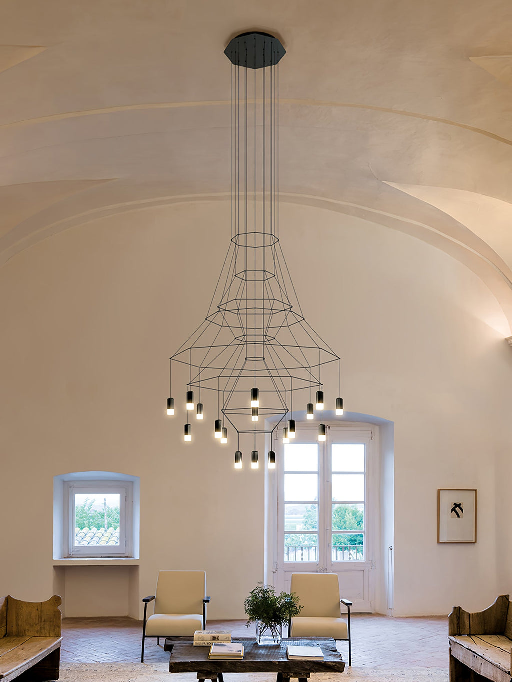 Clérachic wire chandelier with flared frames and a cluster of lit LEDs, hung in a vaulted room with a window and armchairs.