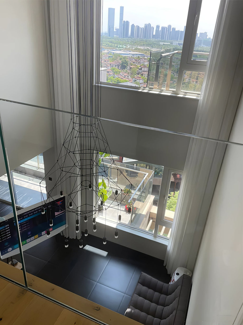 Looking down a stairwell with a glass rail at a Clérachic wire chandelier hung beside tall curtains and a city view.