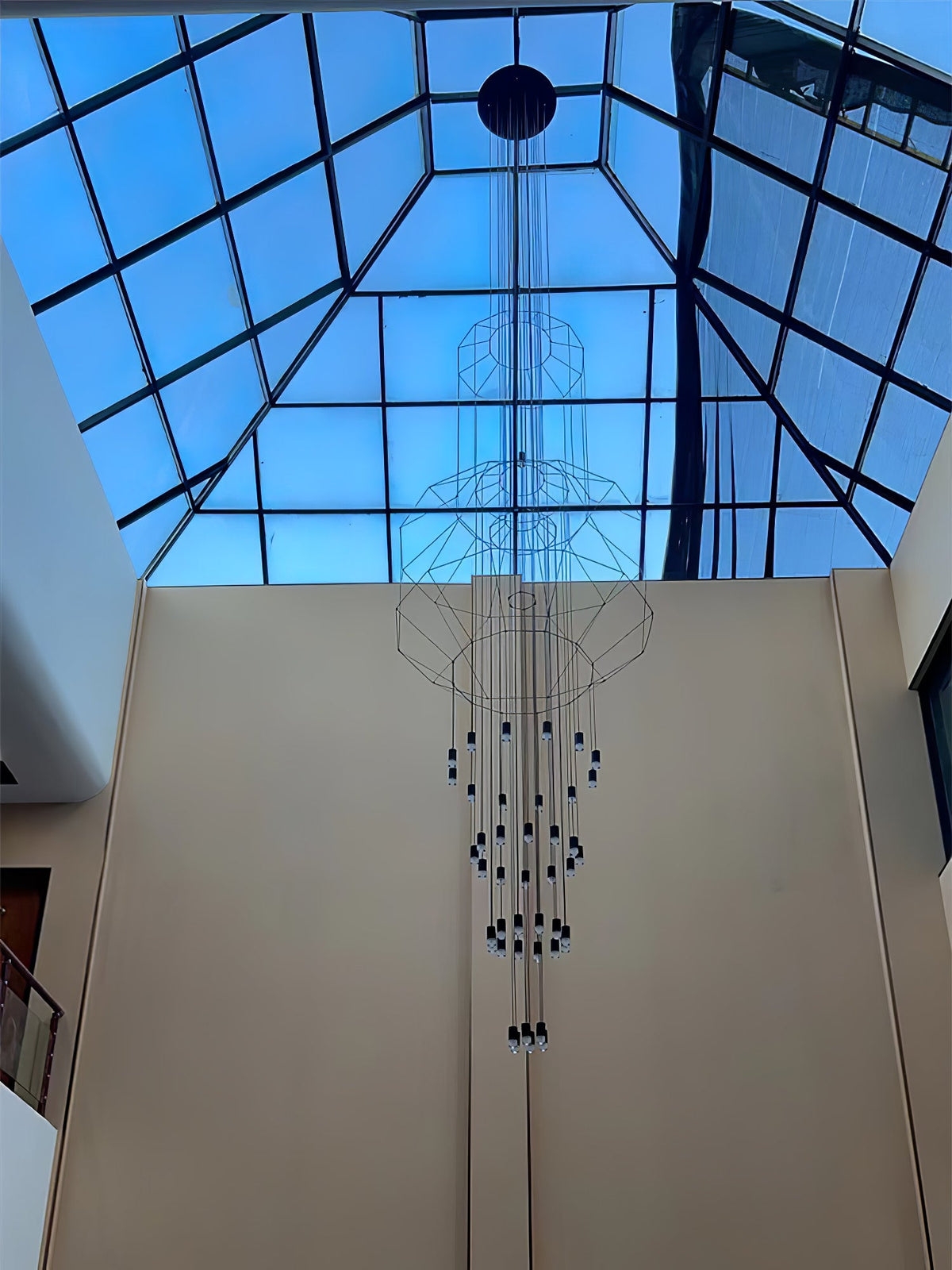 Looking up at a very long Clérachic wire chandelier hung under a glass pyramid skylight against a blue sky.