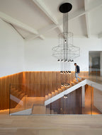A man walks up a timber-clad stair under a hanging Clérachic wire chandelier, with a ceiling of white beams above.