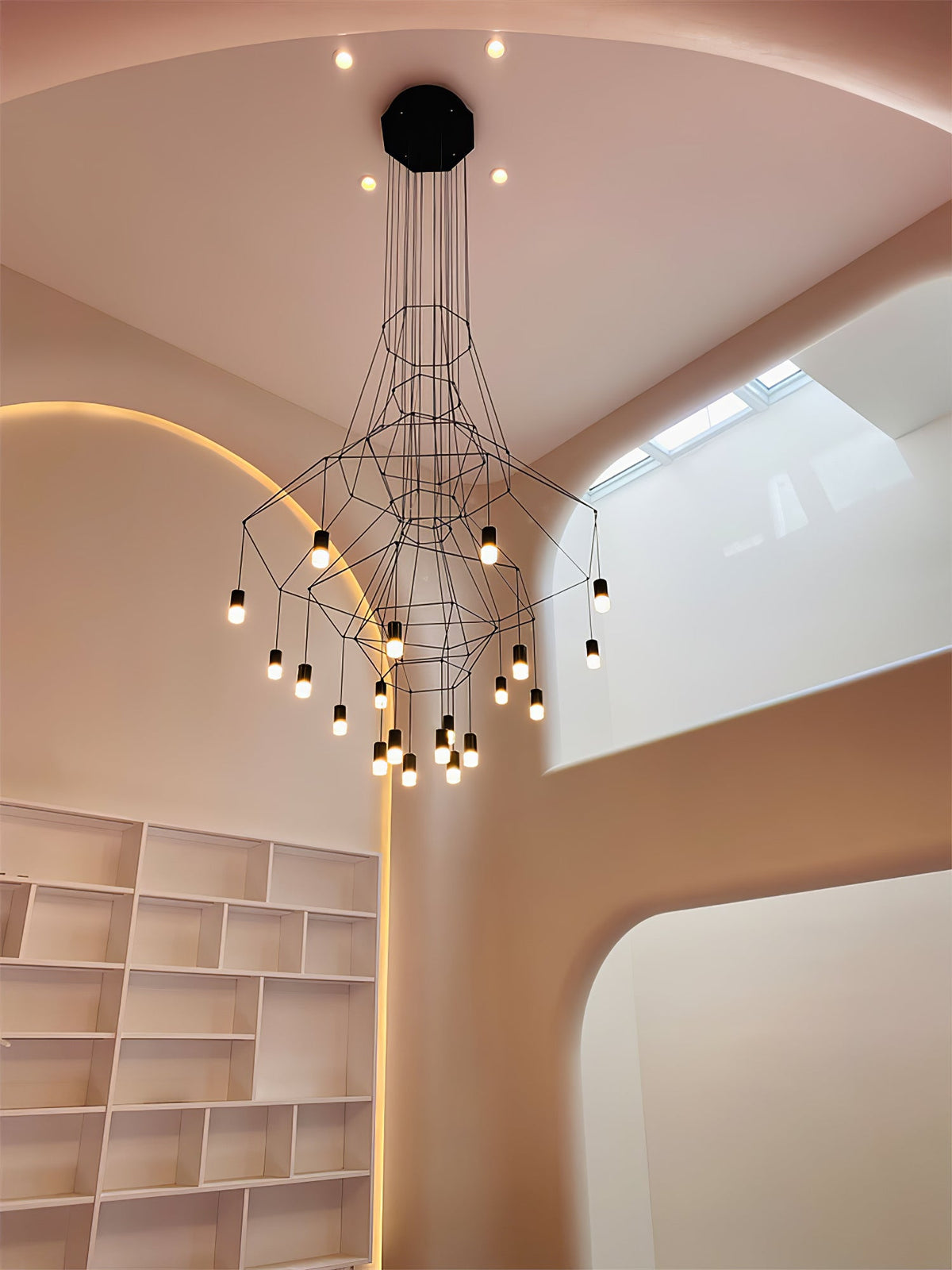 Lit Clérachic wire chandelier hanging by a white bookcase and a curved, softly lit rose-coloured wall.