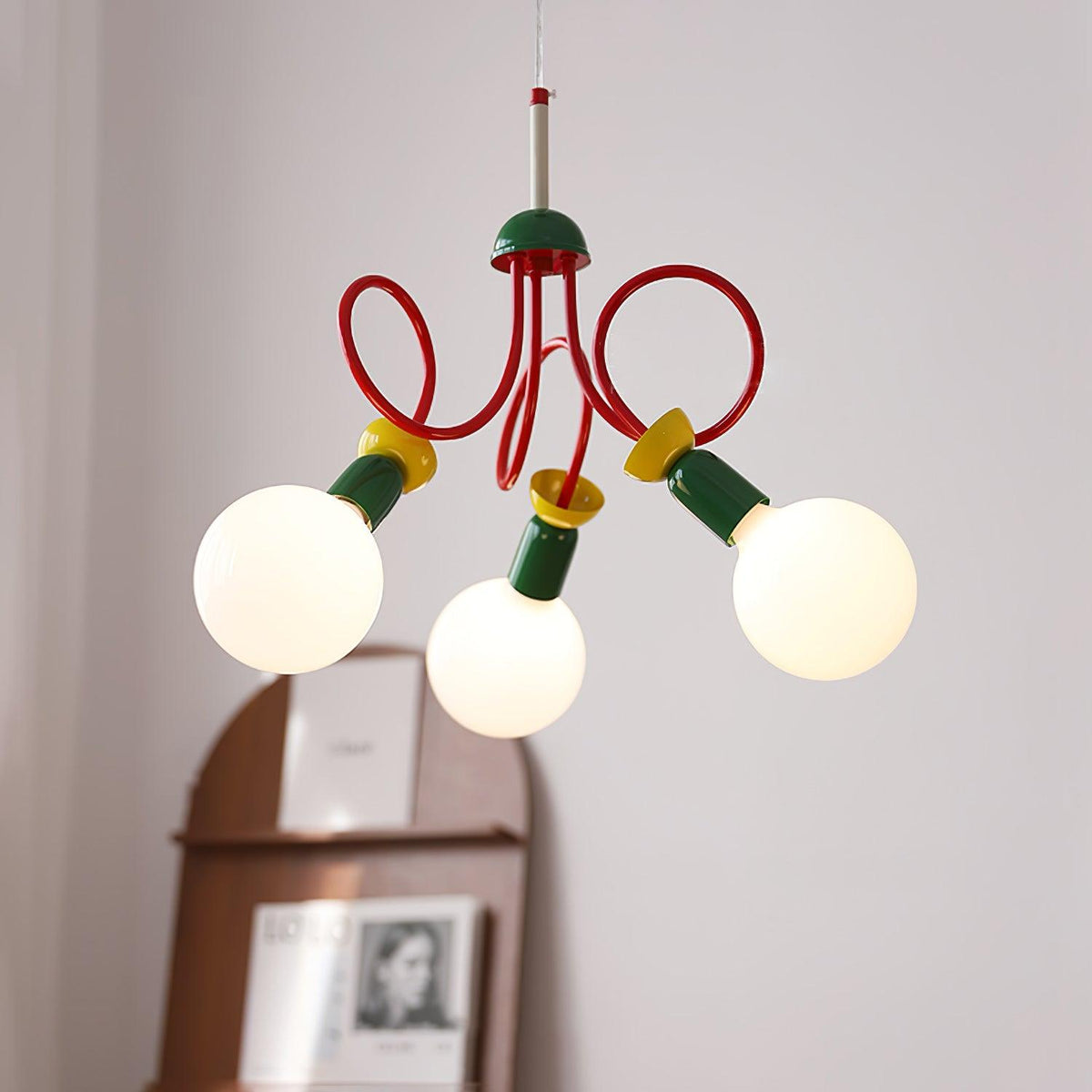 Three globe bulbs on a looping red-armed Clérachic pendant, with an arched wooden shelf and framed print softly out of focus behind.