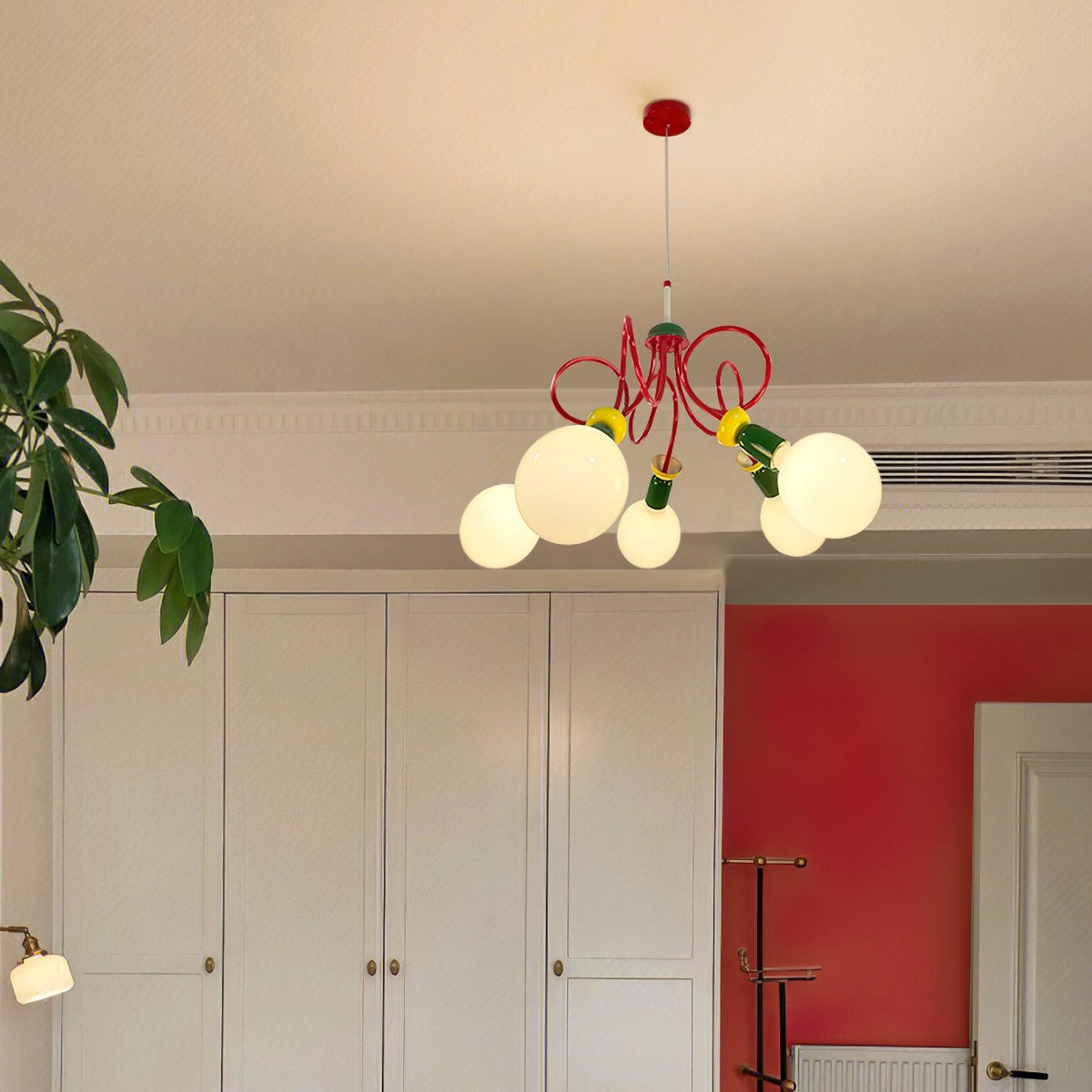 Distant view of this Clérachic pendant glowing high in a room with white cupboards, a red wall and a potted plant.