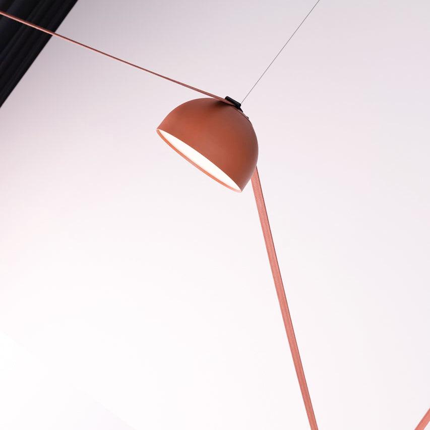 Seen at an angle against a pale pink backdrop, this Clérachic pendant light has an orange dome with a red fabric strap and a thin steel wire.