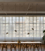 A row of Clérachic pendant lights with dark domes hangs at staggered heights over a long table in a loft with large gridded windows.
