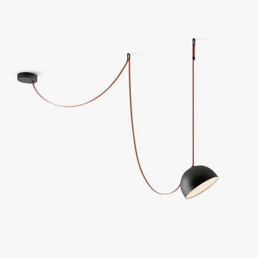 This Clérachic pendant light on a white backdrop has a black metal dome, with its red strap swooping from a round ceiling plate to a wall hook.