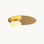Brass Clérachic ceiling light with a milk glass globe set against an oval metal disc, lit on a plain off-white backdrop.