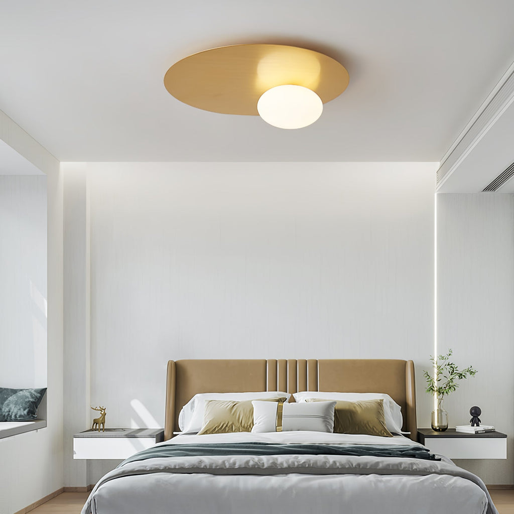A Clérachic brass disc ceiling light with a white glass globe above a tan-headboard bed in a pale, minimal bedroom.
