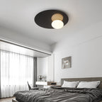 Black Clérachic disc ceiling light with a lit milk glass globe over a bedroom with grey bedding and sheer curtains.