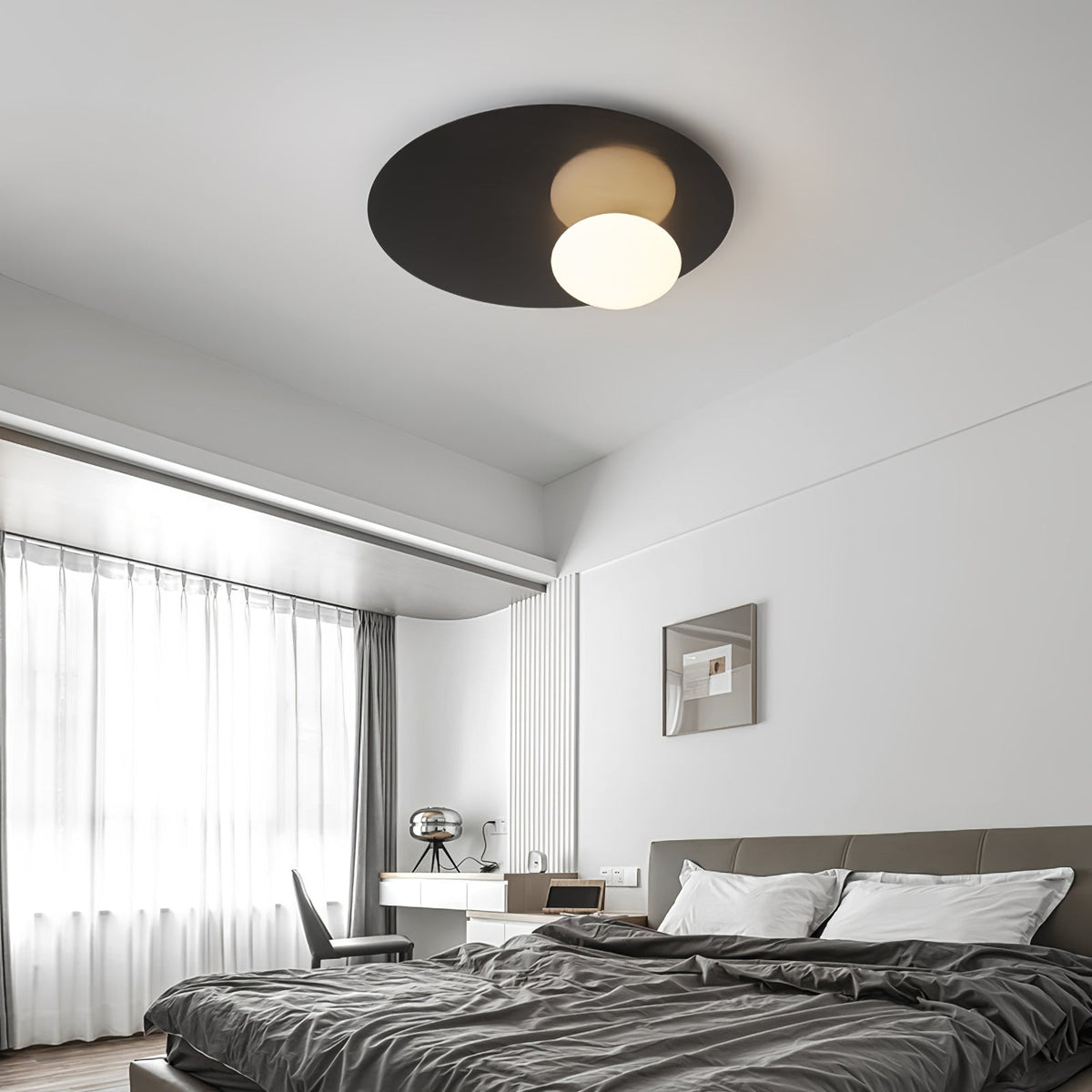 Black Clérachic disc ceiling light with a lit milk glass globe over a bedroom with grey bedding and sheer curtains.