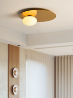 Brass Clérachic ceiling light lit beside a ceiling recess, with pleated curtains and a wood panel in the background.