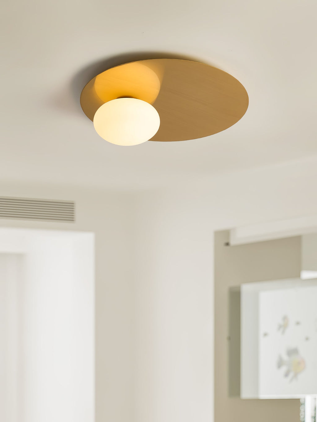 Lit brass Clérachic ceiling light with a glass globe, seen from below in a bright room with a slatted blind.