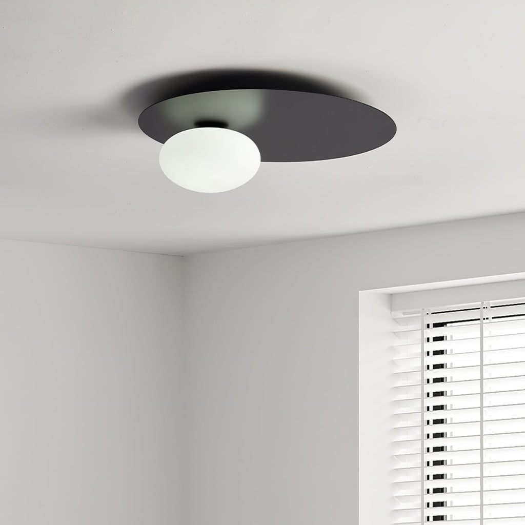 Black Clérachic disc ceiling light with an unlit white glass globe, seen at the corner of a room near a window blind.