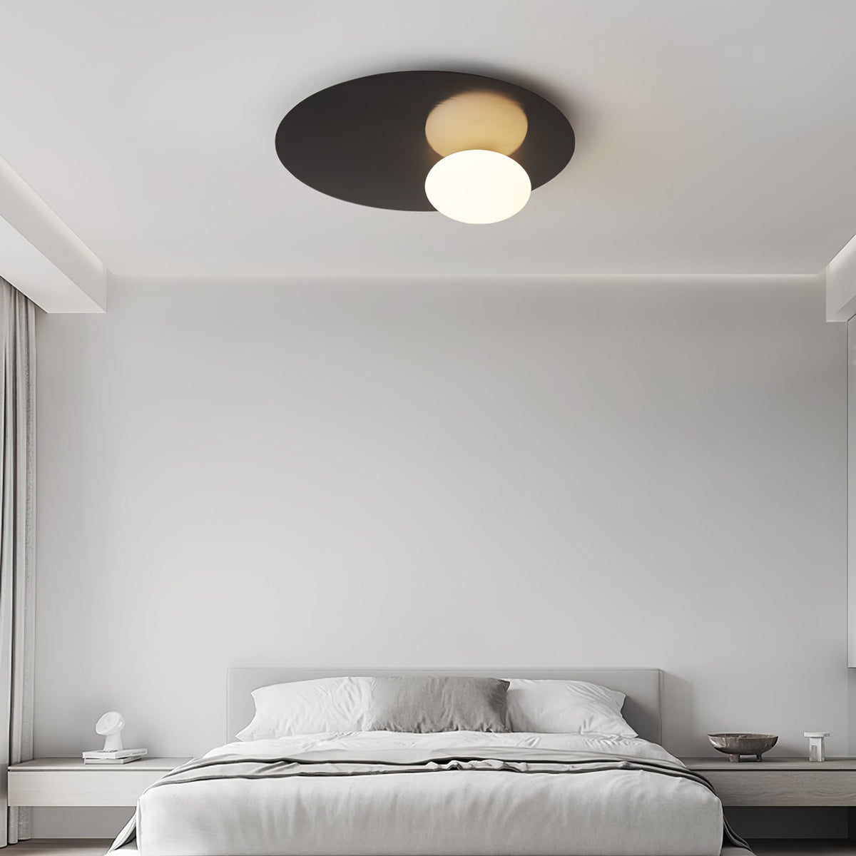 Black Clérachic disc ceiling light with a lit globe above a neatly made bed in a quiet grey bedroom.