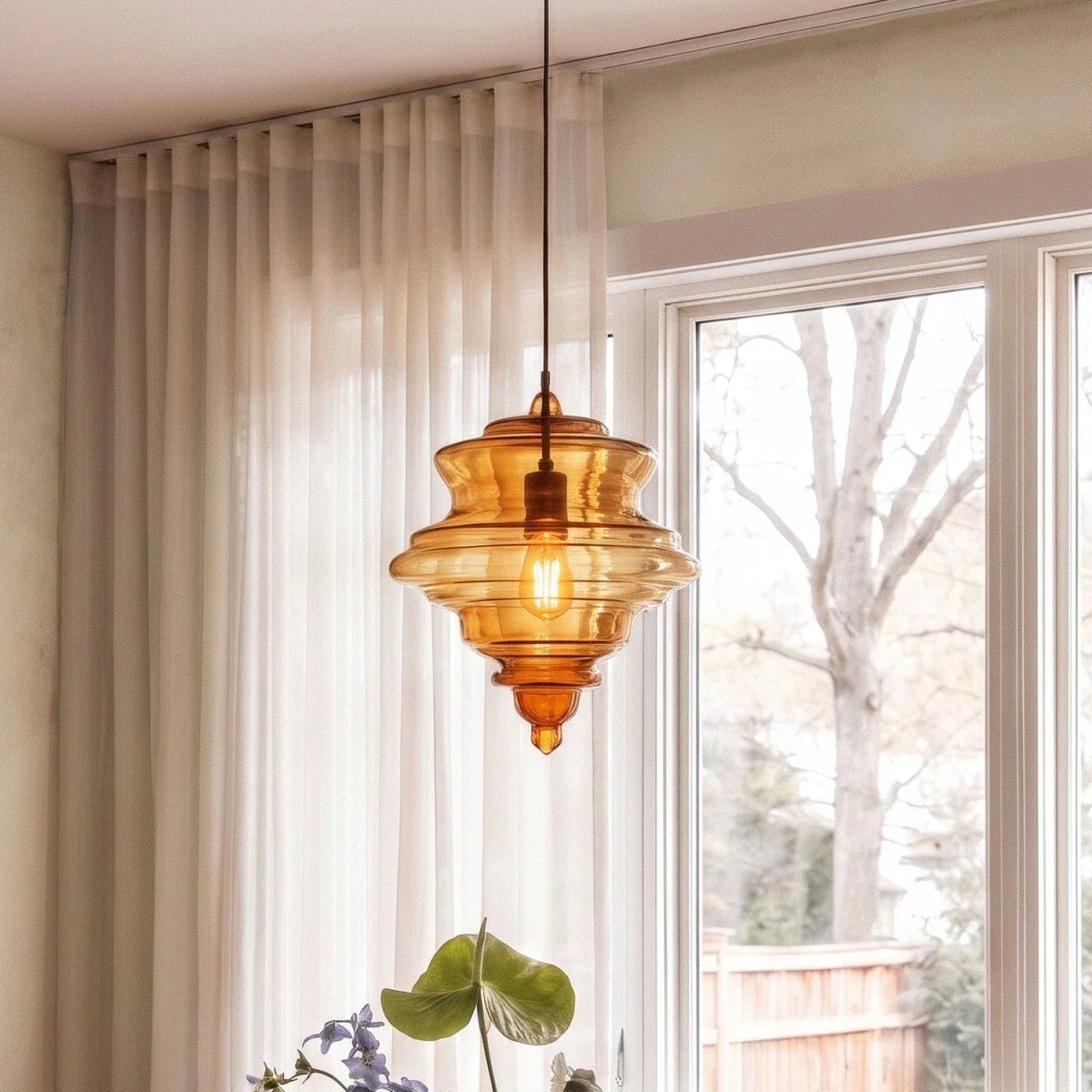 A closer view of the Clérachic pendant light in cognac glass, glowing warm beside a sheer curtained window.