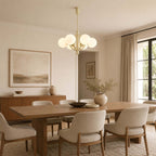 A Clérachic chandelier with white glass globes hangs over a long walnut dining table with upholstered chairs in a pale, calm room.
