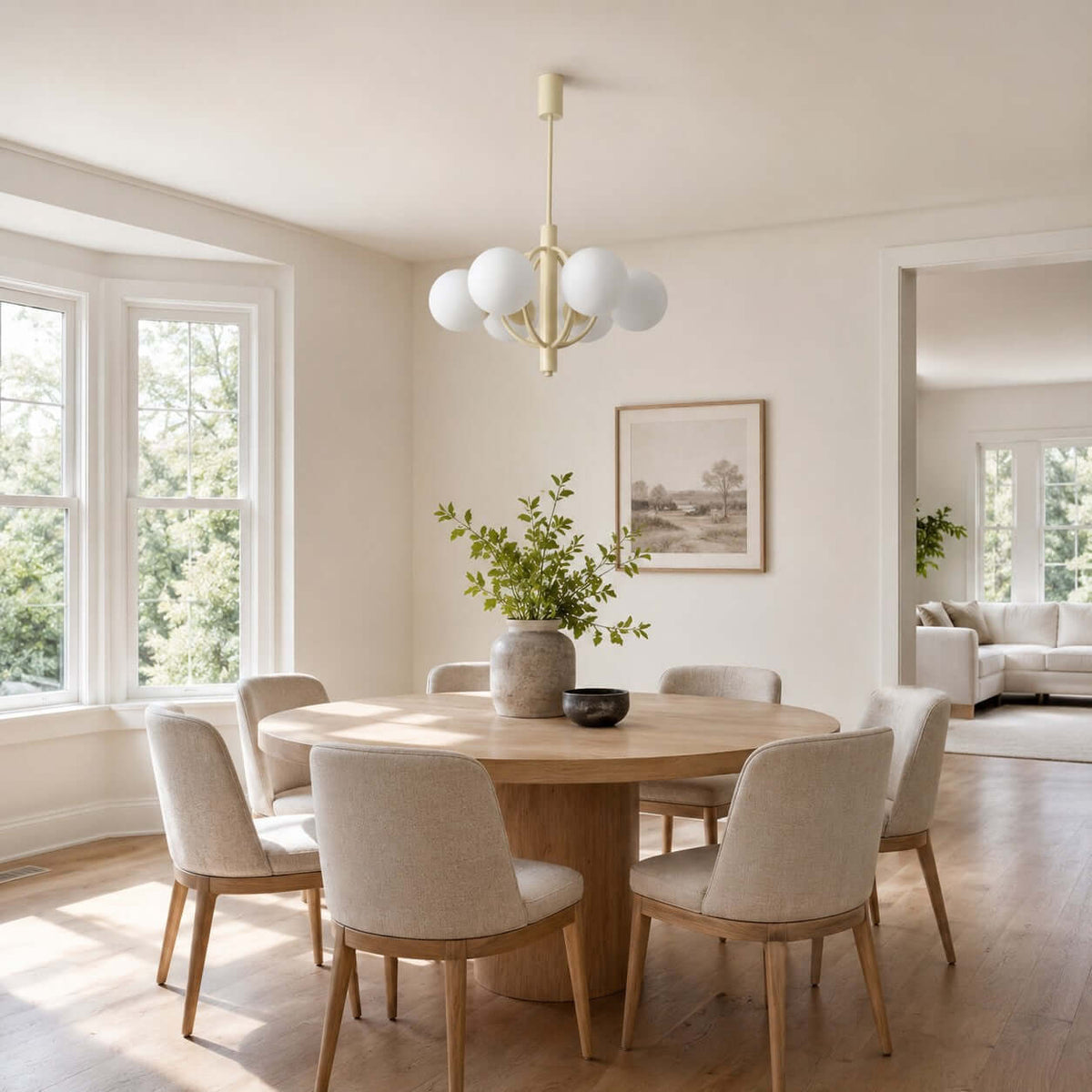 A Clérachic chandelier hangs over a round pale-wood dining table near a bay window with a leafy vase on the table.