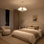A Clérachic chandelier glows warm above a bed with caramel bedding, with a bouclé armchair and a dark night window.