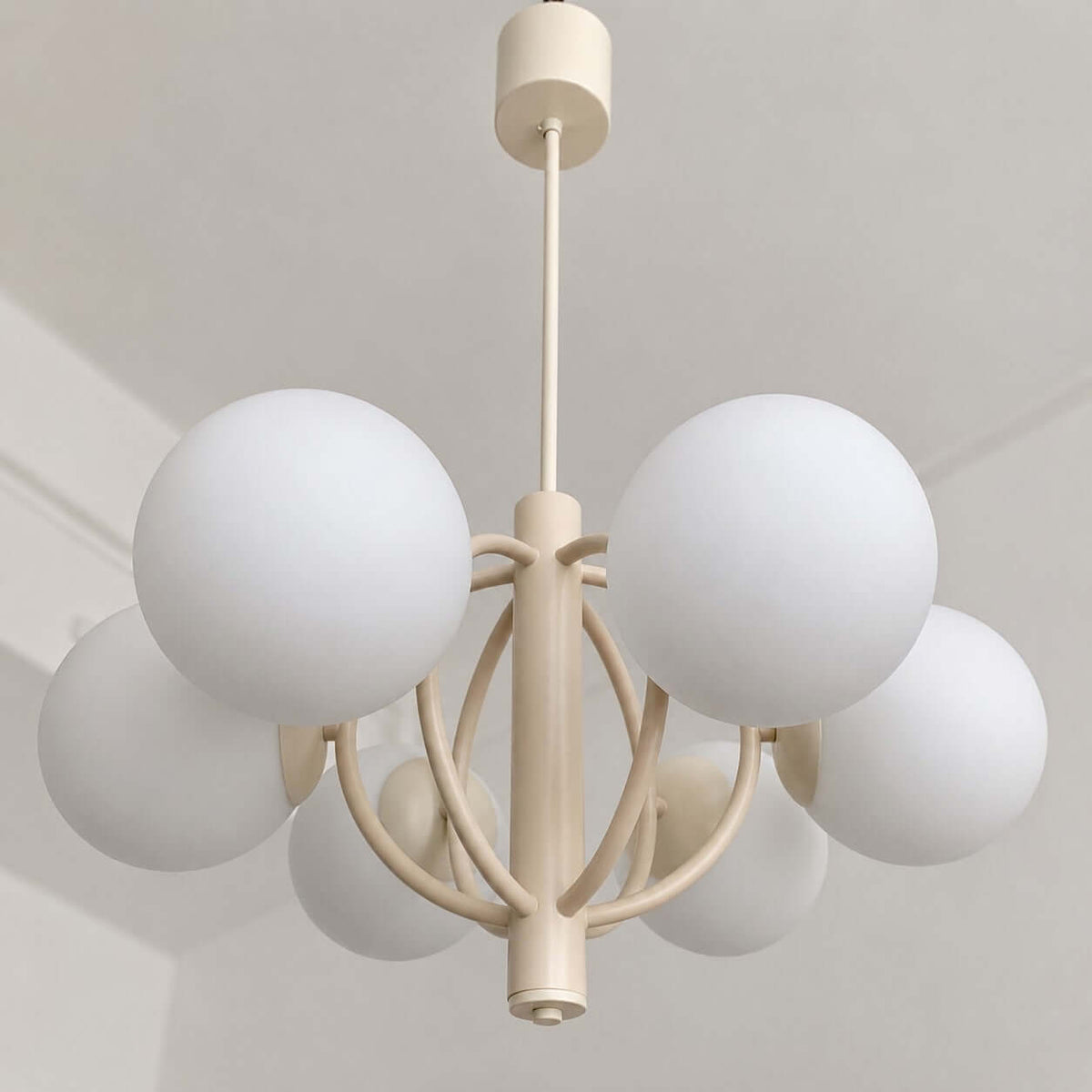 Looking up at a Clérachic chandelier, with its white glass spheres, curved beige arms and the ceiling canopy above.