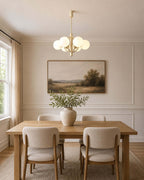 A Clérachic chandelier hangs over a rectangular wooden table, with a landscape painting on a panelled wall behind.