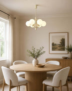 A Clérachic chandelier with white globes, lit, above a round pedestal dining table in a bright room with a landscape print.