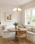 A Clérachic chandelier hangs in a breakfast nook with a round table, a bench seat and a window with a leafy view.
